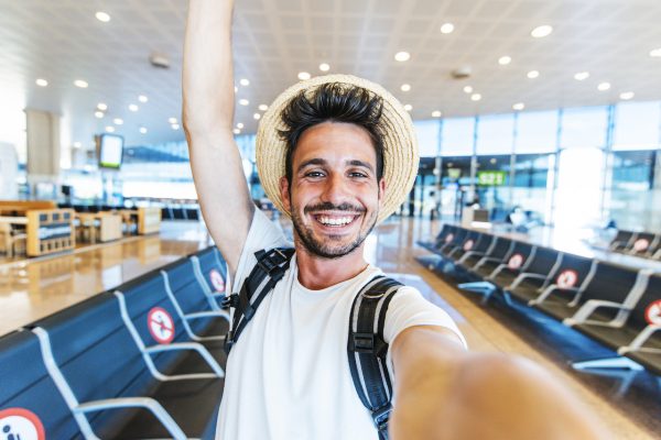 Happy tourist man taking a selfie portrait with mobile smart phone at airport - Airplane passenger waiting for flight - Travel without restrictions concept